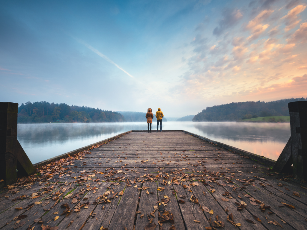 Two people standing on a dock, looking over a lake during the sunset.